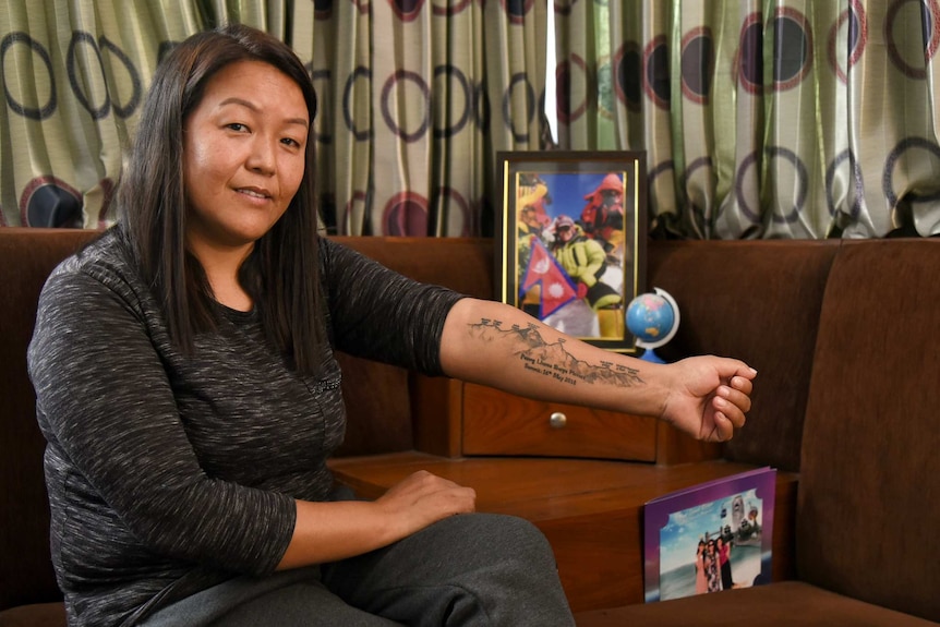 A woman with brown skin and hair wearing a black top sits down as she shows off a Mount Everest shaped tattoo on her arm.