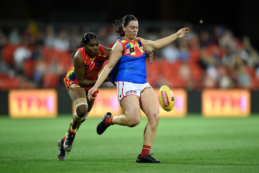 A Melbourne AFLW player drops the ball to kick it downfield as a Gold Coast Suns player tries to tackle her from behind.