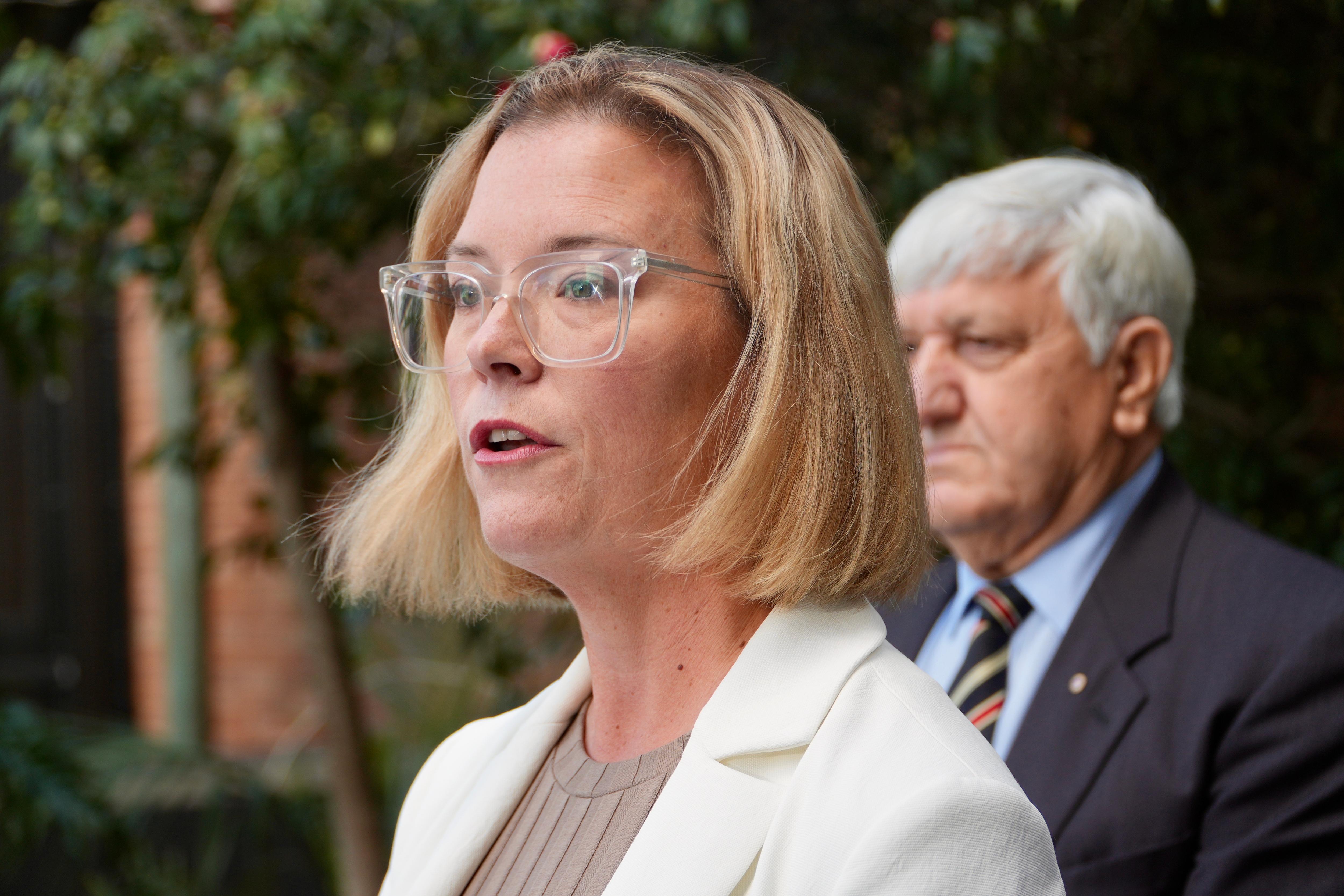 A blonde woman wears spectacles and a light-coloured blazer as she stands outdoors and speaks.
