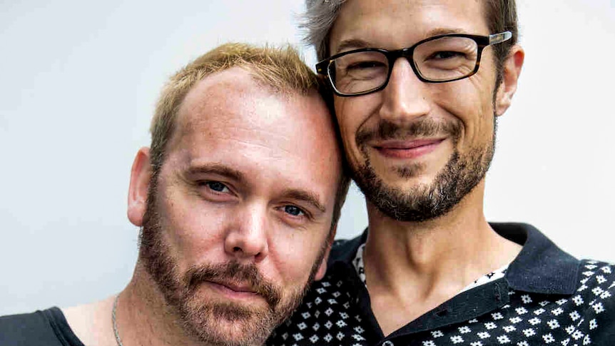 Man with blond hair and beard leans on man with glasses, smiling with his arm around his partner.