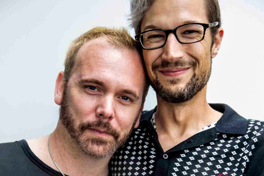 Man with blond hair and beard leans on man with glasses, smiling with his arm around his partner.