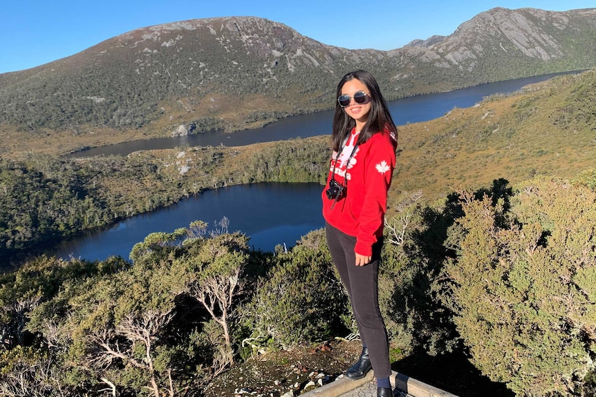 A woman in a red jumper stands in front of a natural vista featuring a river and mountains.