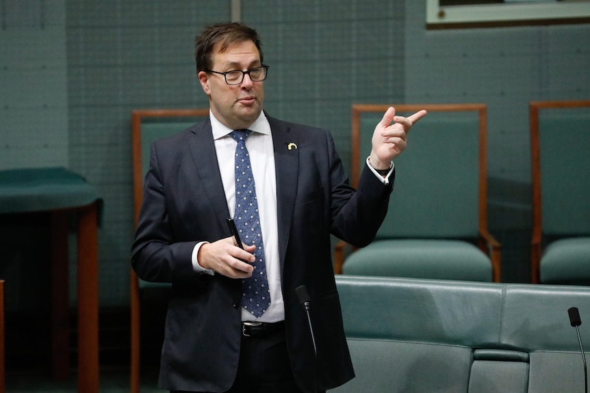 Falinski wearing a dark suit and glasses, holds the phone in his right hand, points with his left.