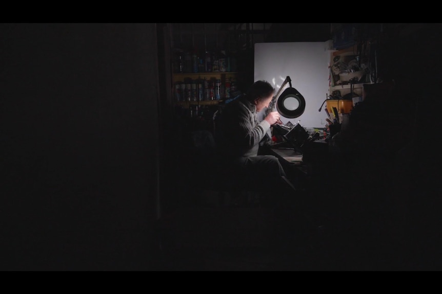 A man sits silhouetted at his desk, repairing a typewriter.