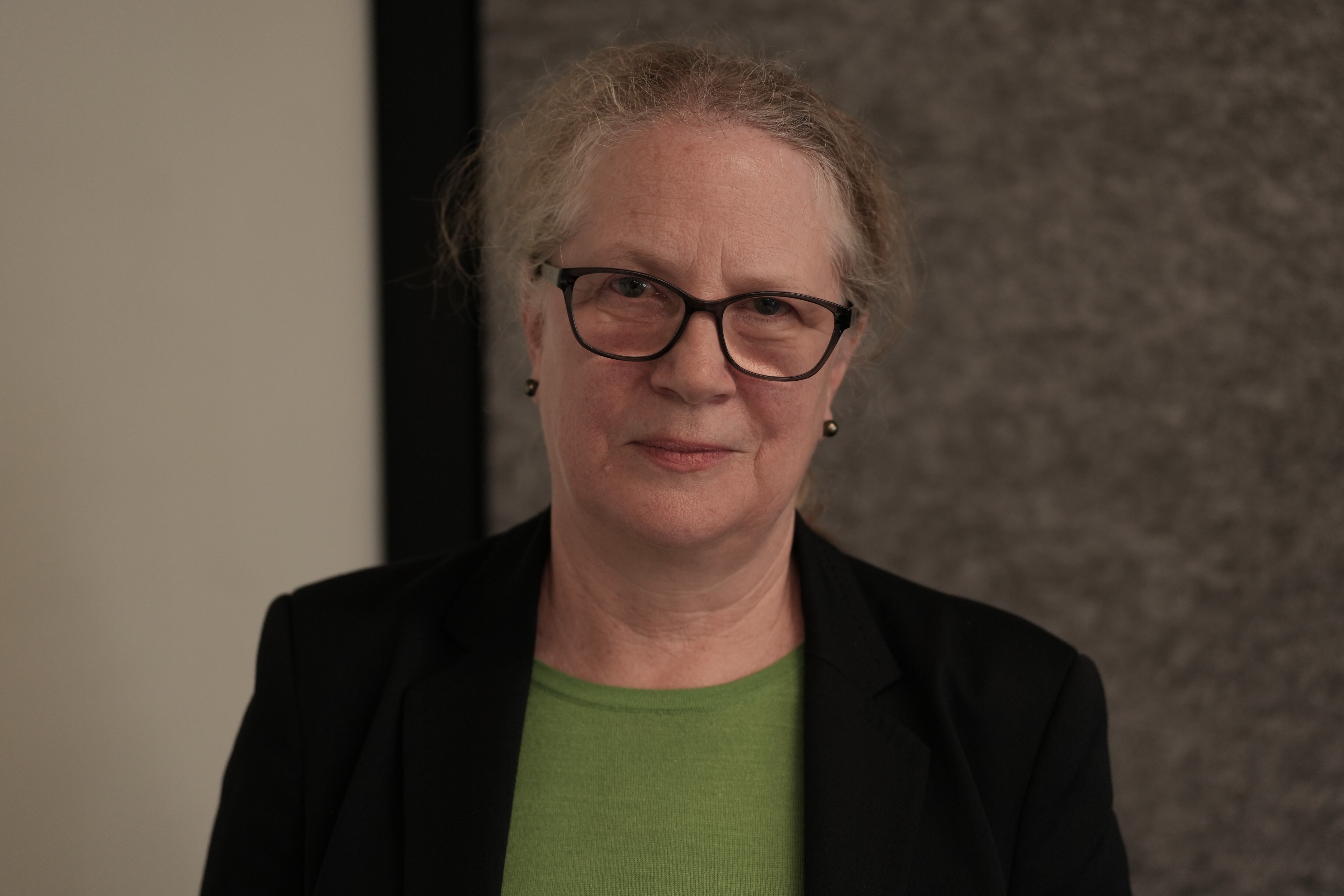 A woman with light curly hair and glasses stands indoors looking serious.