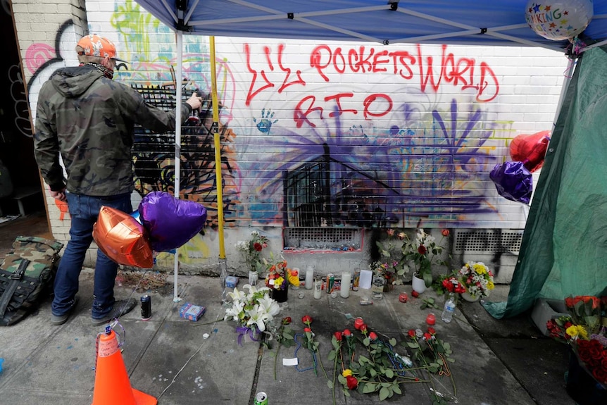 a man wearing a mask spray paints a wall at a memorial that includes balloons and flowers