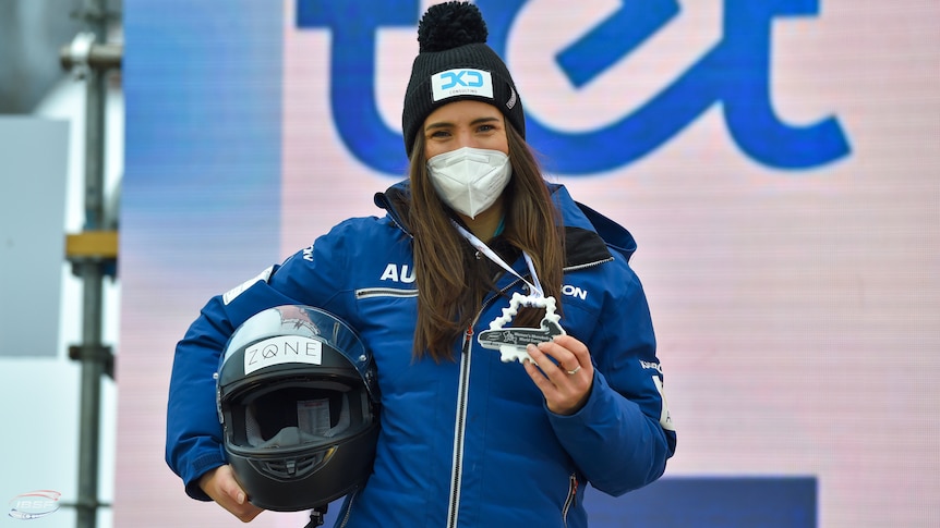 Bree Walker, wearing a mask, holds a helmet under one arm and a medal in the other