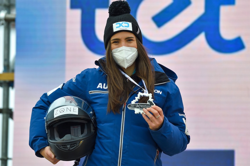 Bree Walker, wearing a mask, holds a helmet under one arm and a medal in the other