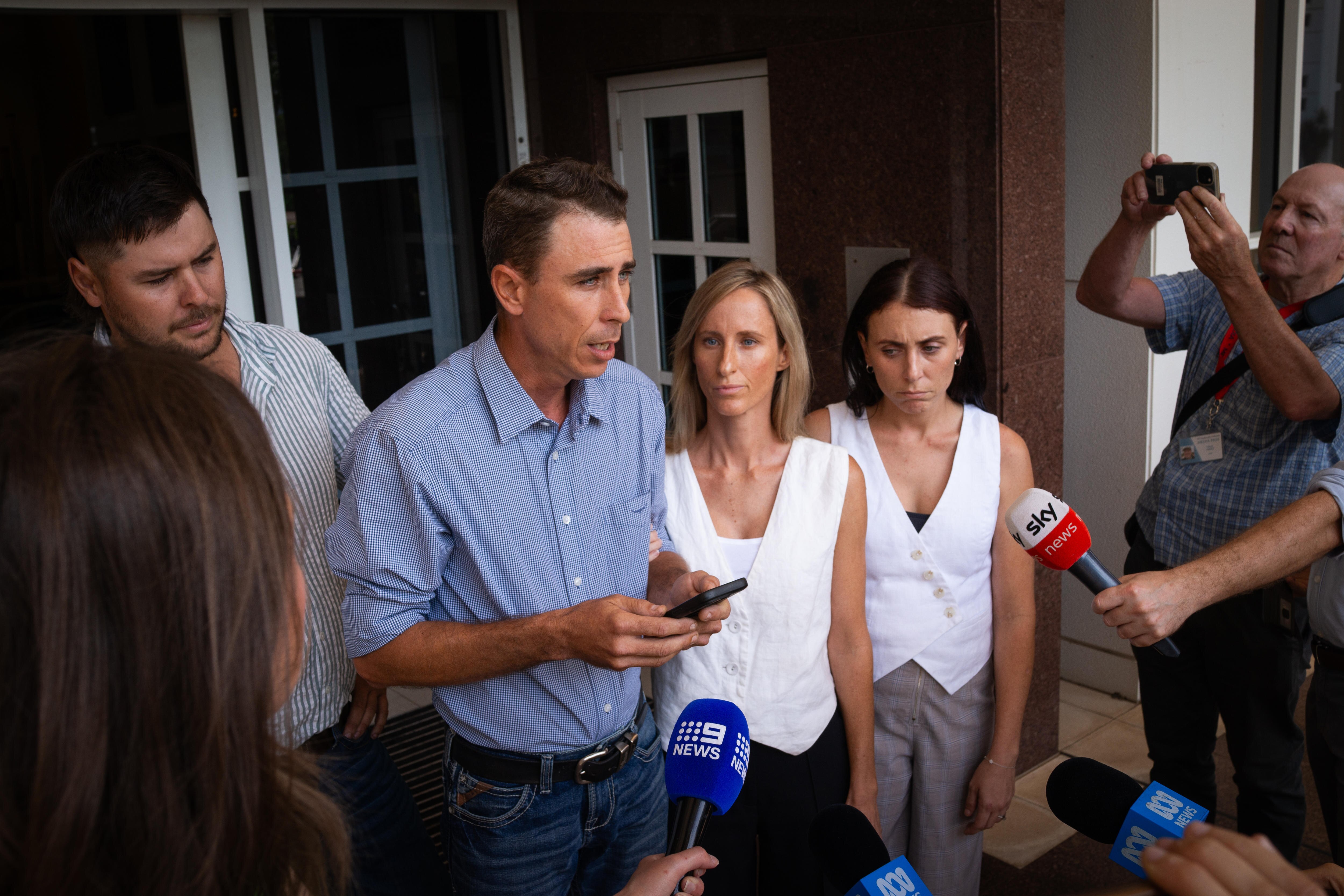 A man in a button-up shirt speaking in front of news microphones, with family members at his side.