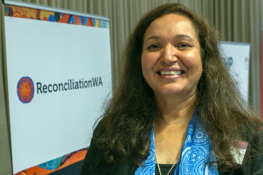 A woman in a black top and a bright blue scarf stands in front of a Reconciliation WA sign.