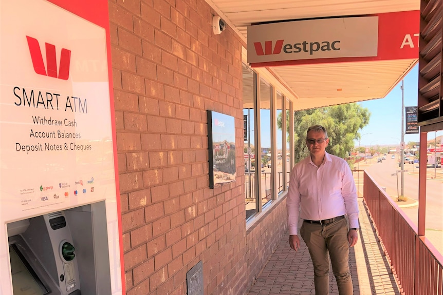A serious older man wearing pink shirt and brown trousers, grey hair, stands in a brick verandah with Westpac sign and ATM. 