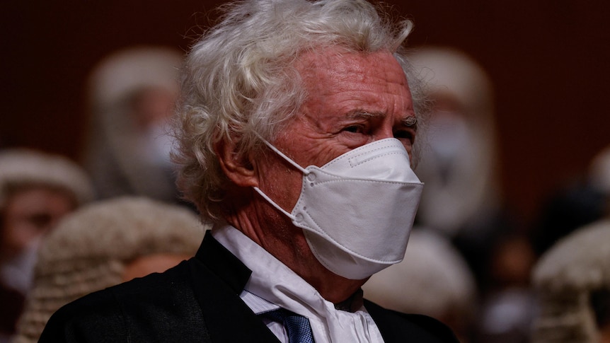 A man with grey curly hair wears a white face mask.