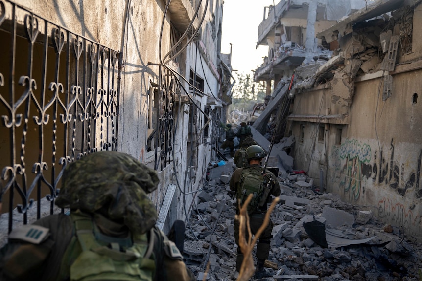 soldiers walk through an alleyway between buildings, rubble and debris lining the ground, one of the buildings is badly damaged