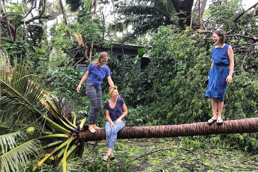 Three women balance on a fallen palm tree, two standing while the other sits. The background is green with debris.