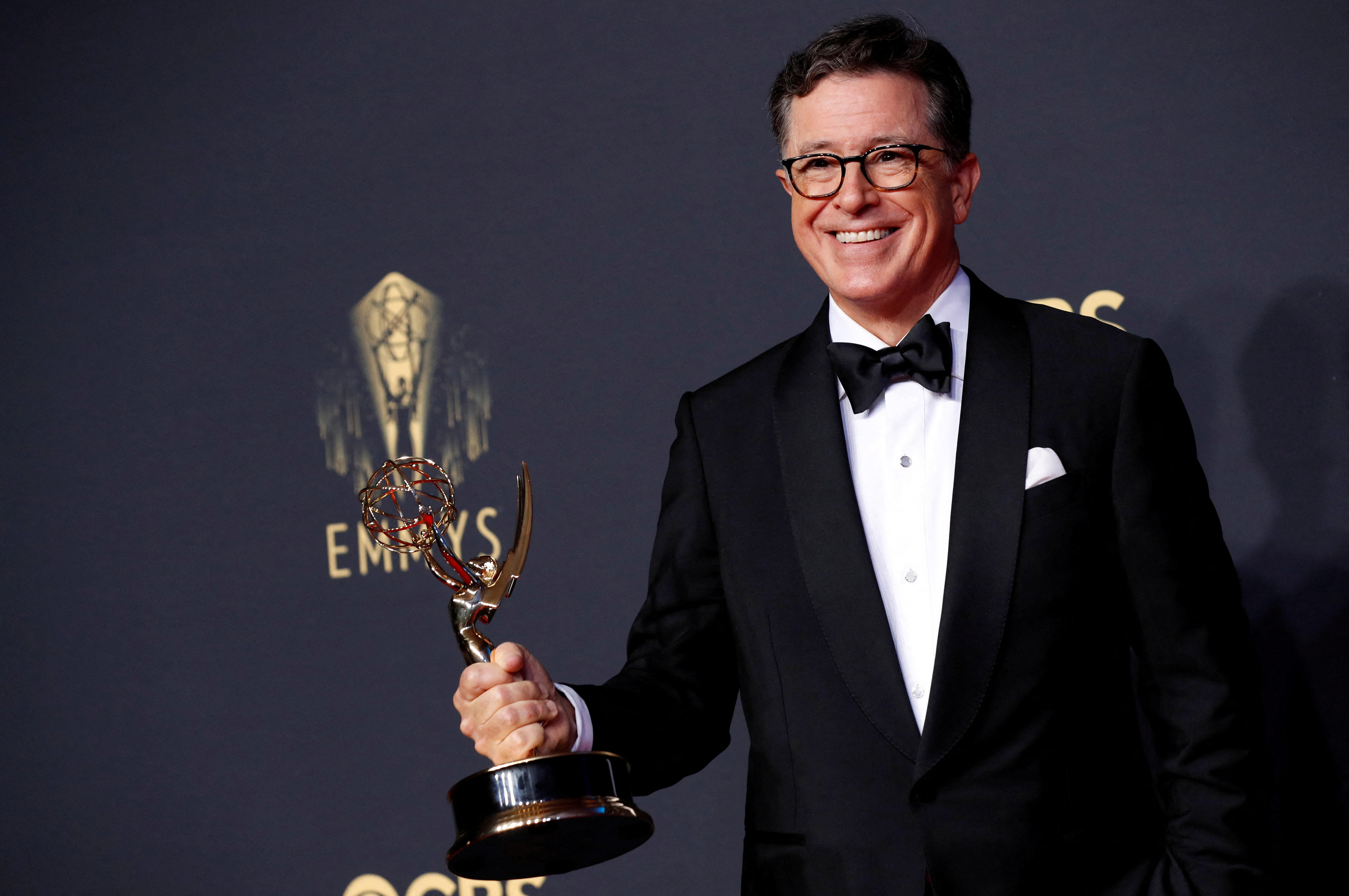 A middle-aged man in a tuxedo and glasses beams as he holds a golden Emmy award trophy.