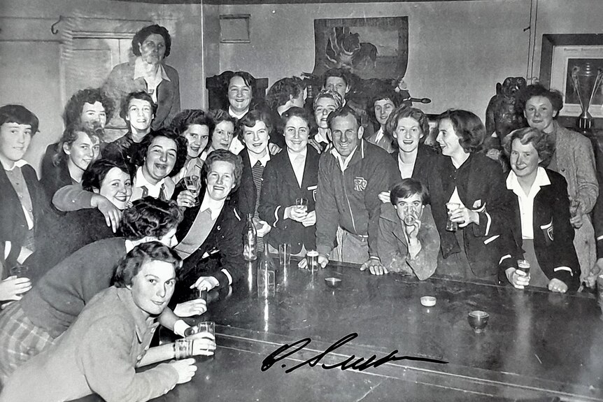 A group of young women gathered around a table with a middle aged man in a football club room