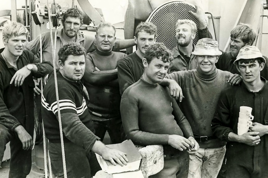 Black and white photo of a group of 10 male naval personnel on a boat, two of them in wetsuits and two wearing bucket hats