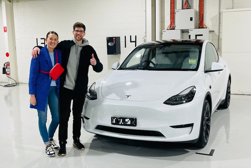 A man and a woman stand next to a white Tesla Model Y car in front of a brick wall painted cream.