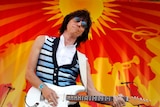 Dark-haired guitarist plays in front of a red and yellow curtain with the image of a rock band on it