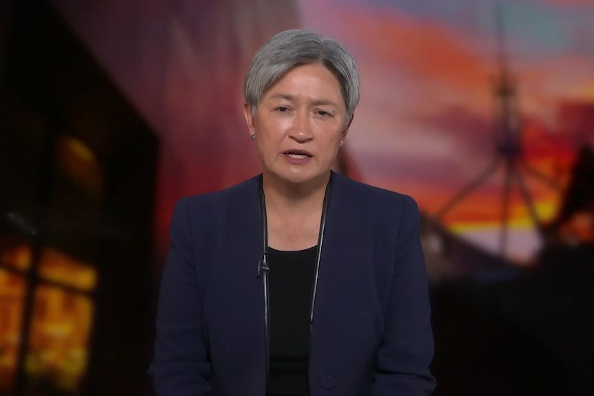 Screen shot of Penny Wong with short grey hair and navy blazer in front of parliament house back drop at sunrise. 