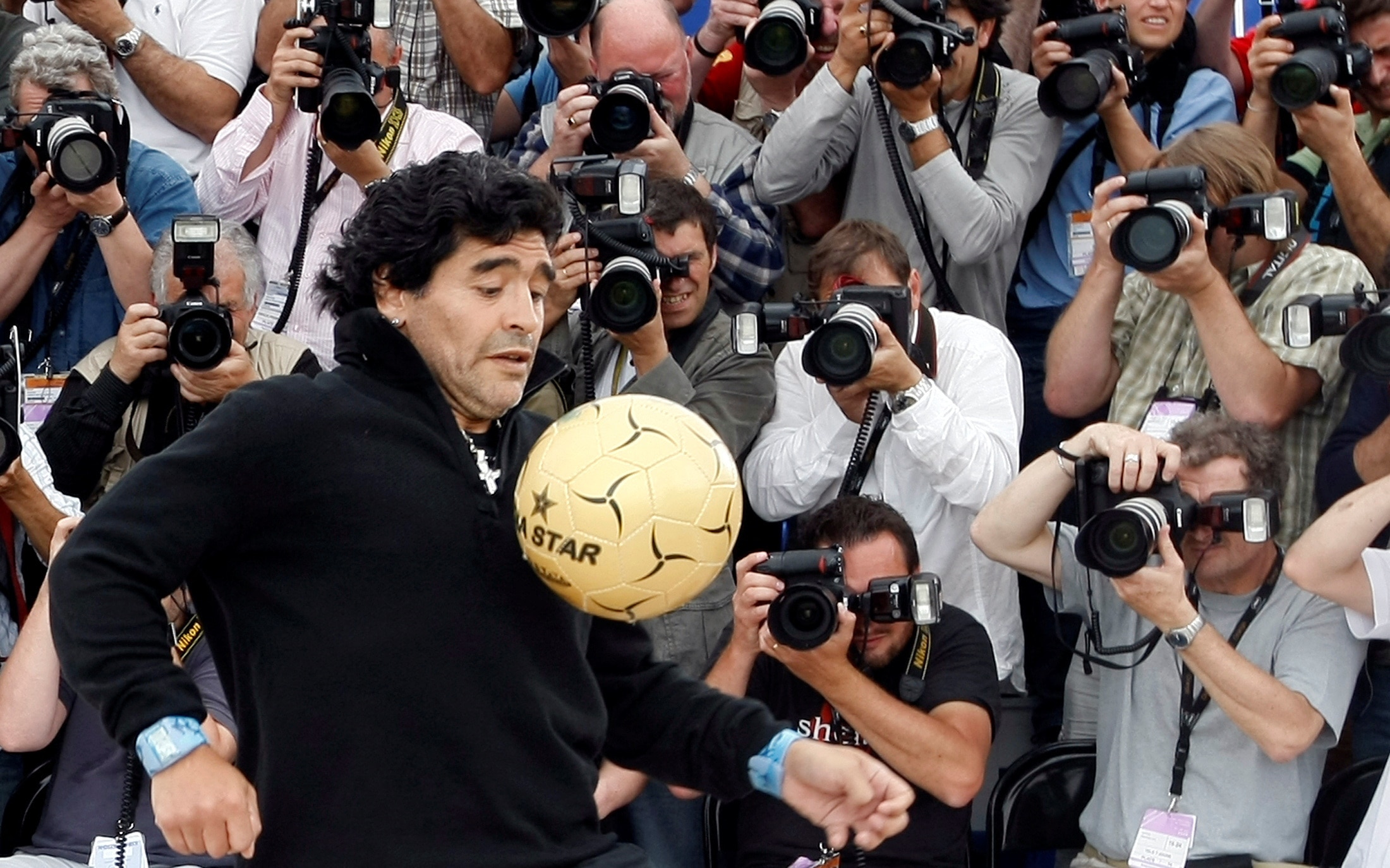 A man bouncing a soccer ball in front of several photographers. 