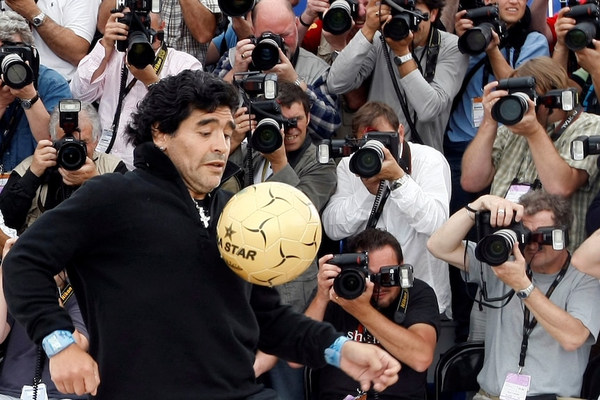 A man bouncing a soccer ball in front of several photographers. 