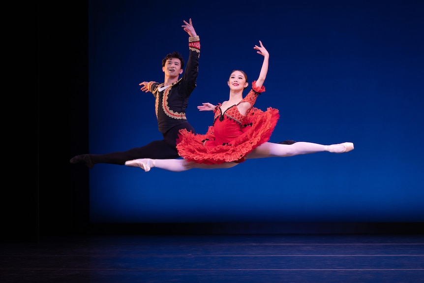A man and a women jump in the air, doing the splits, with one arm in the air