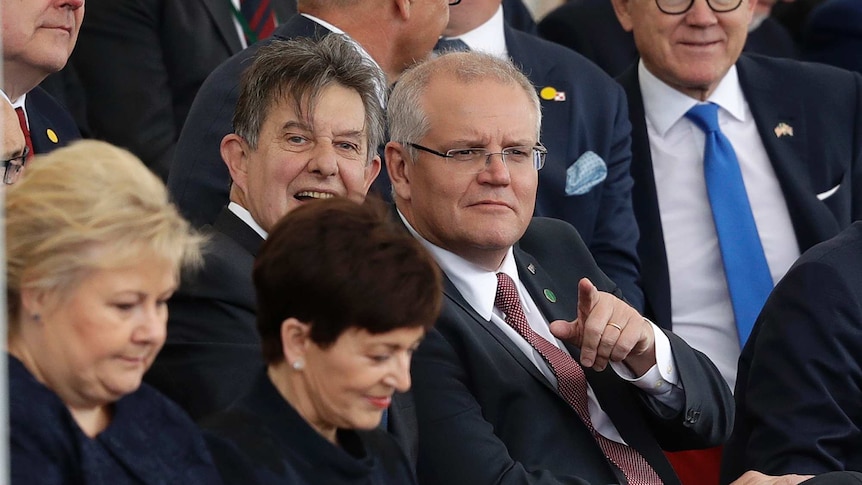 Scott Morrison sits in a crowd of suits and points into the distance.