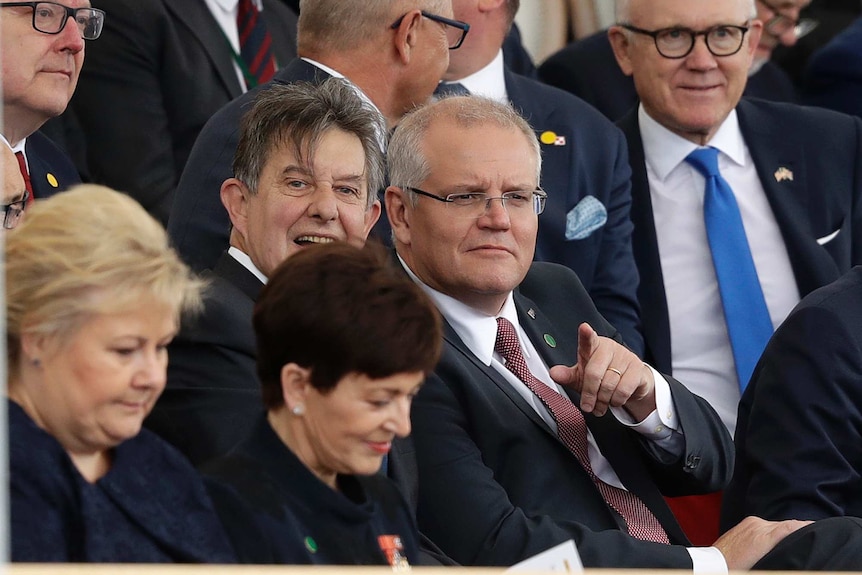 Scott Morrison sits in a crowd of suits and points into the distance.