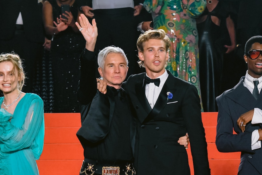 Middle-aged white man with silver hair wears black tuxedo on red carpet beside young white man with blonde hair and black tuxedo