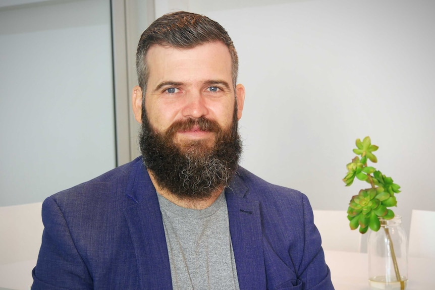 A man with a beard sits in a white board room next to a succulent in a glass jar.