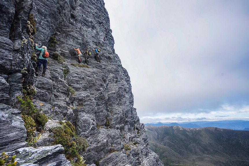 three people a climbing a sheer rock face on a mountain, holding on to the side