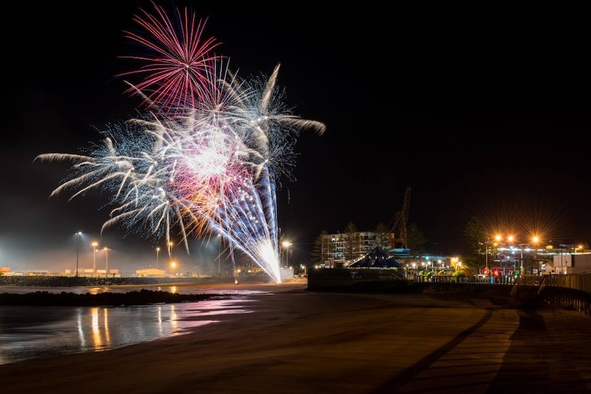 Fireworks over a dark beach with city lights in the background.