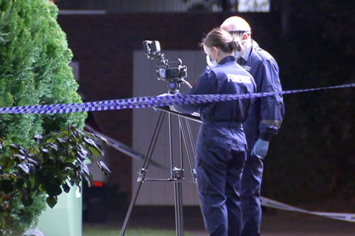 A young female and an older male forensic police officer, dressed in overalls, photograph a crime scene behind police tape.