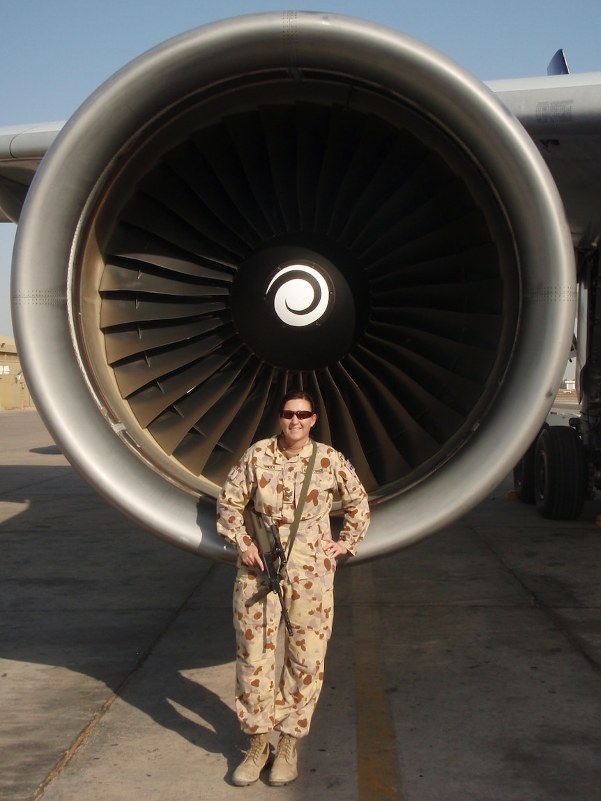 A woman in camouflage holds a gun and smiles for the camera in front of a large aircraft.