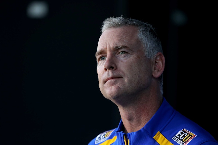 Adam Simpson looks into the distance wearing a blue polo shirt in front of a dark background