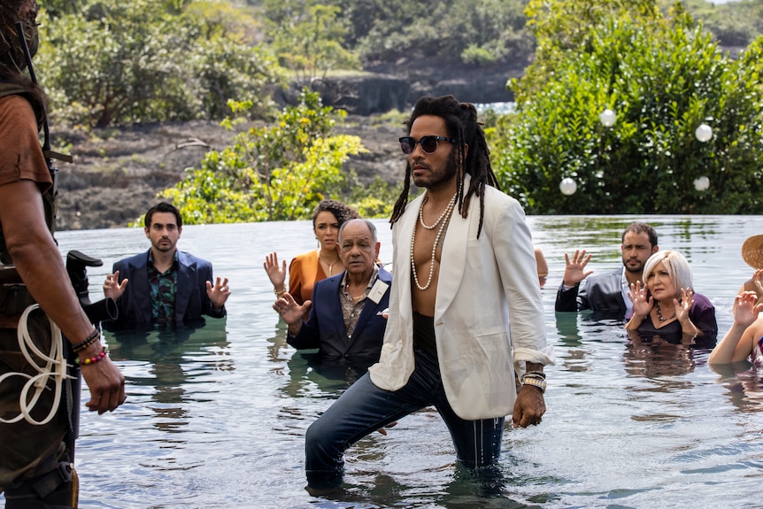 Black man with dark dreadlocks wearing a white blazer stands in shallow river with several people raising arms in surrender.