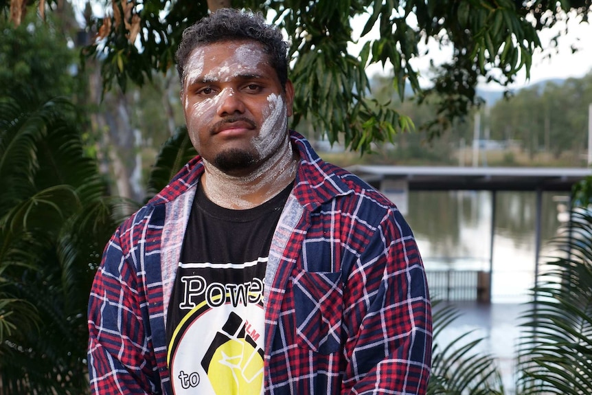 Matthius Mann stands on the Rockhampton riverbank with white paint on his face and a serious expression.