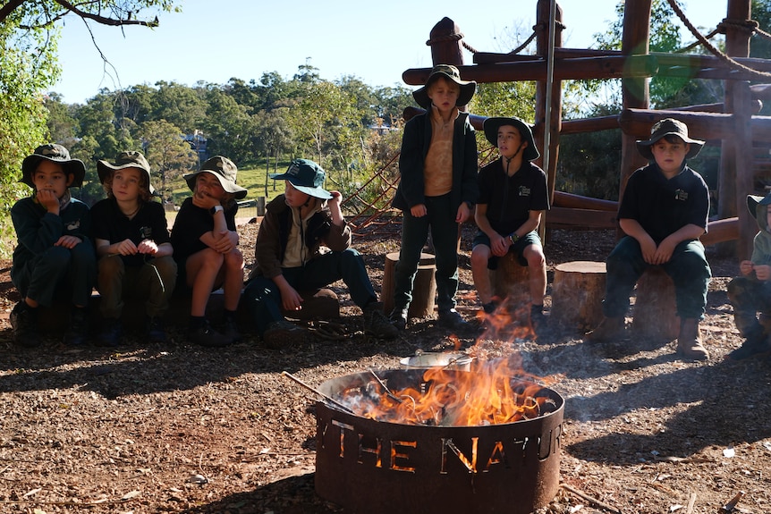 A group of primary school boys sit around a fire pit wearing green hats and uniforms.