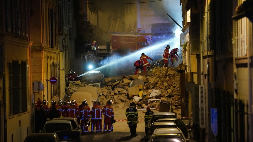 Firefighters fire a stream of water diagonally up into a collapsed building while standing on rubble.