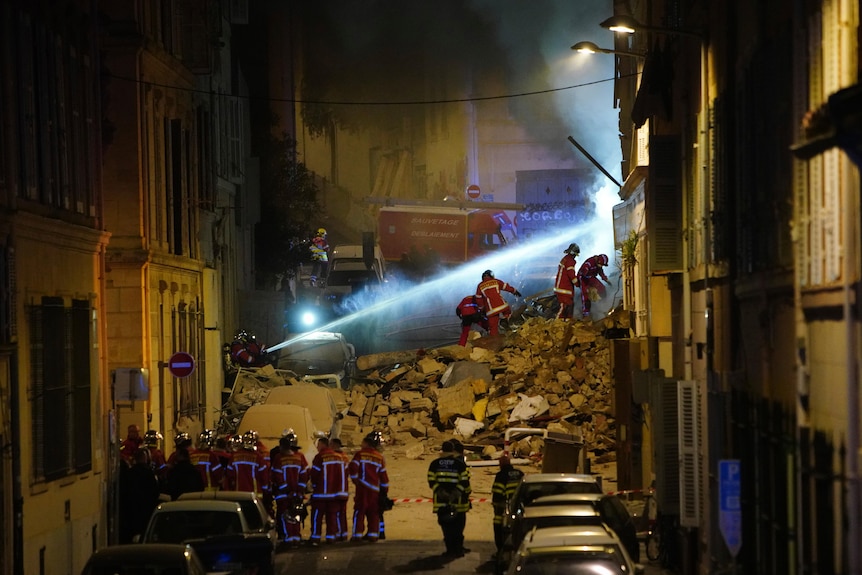 Firefighters fire a stream of water diagonally up into a collapsed building while standing on rubble.