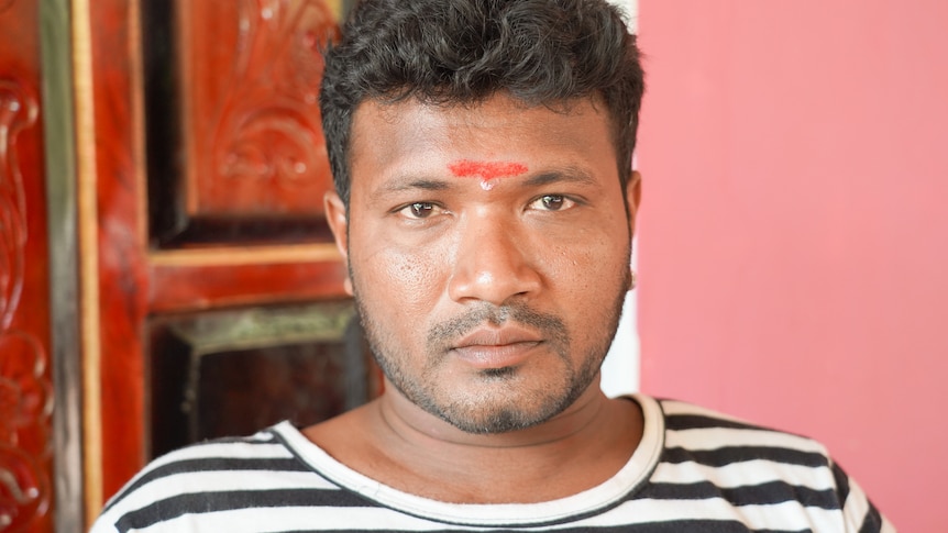 A man wearing a striped shirt with red paint inbetween his eyebrows stands infront of a door.