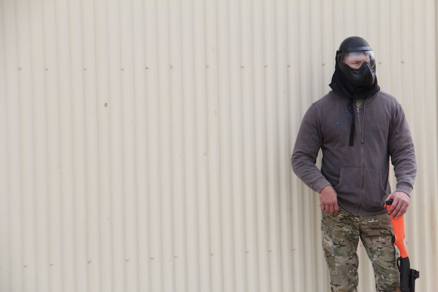 A man in a mask leans on a fake fun against a shed wall.