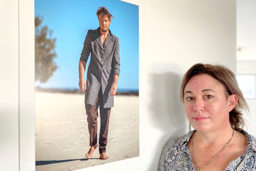 A woman looks into the camera while standing beside a photograph