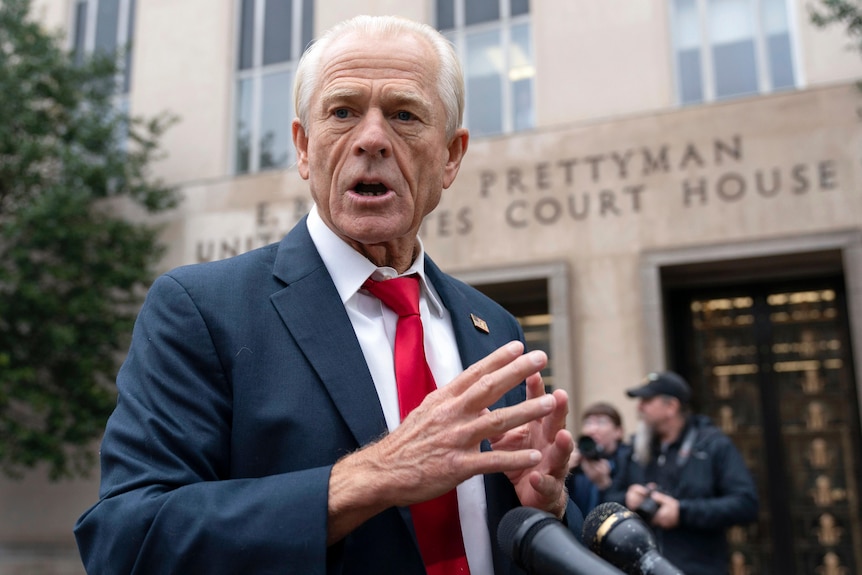 A close up of an elderly man in a suit and tie speaking outside a court building into microphones and gesturing with his hands