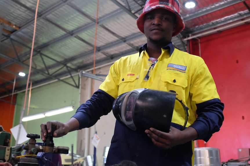 A man in hi-vis overalls stands holding a welders helmet.