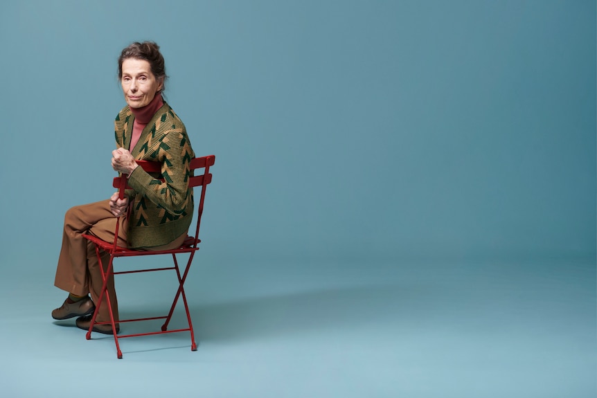 An older woman sits on a fold-out chair, which is facing away from the camera. She turns, an arm over its back, to face the lens