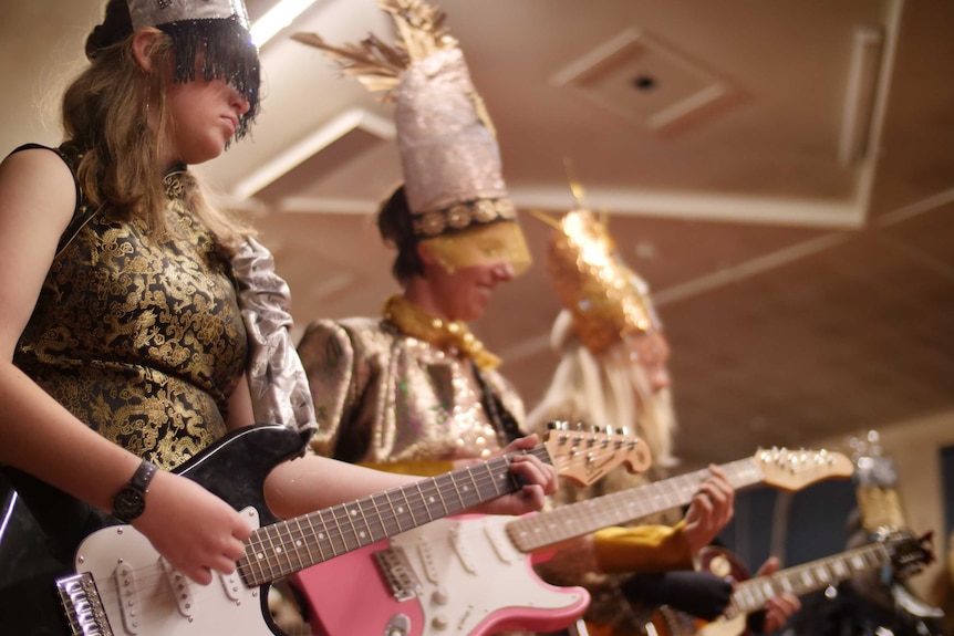 Three women playing guitar while dressed up in gold, black and silver costumes, including tall headresses