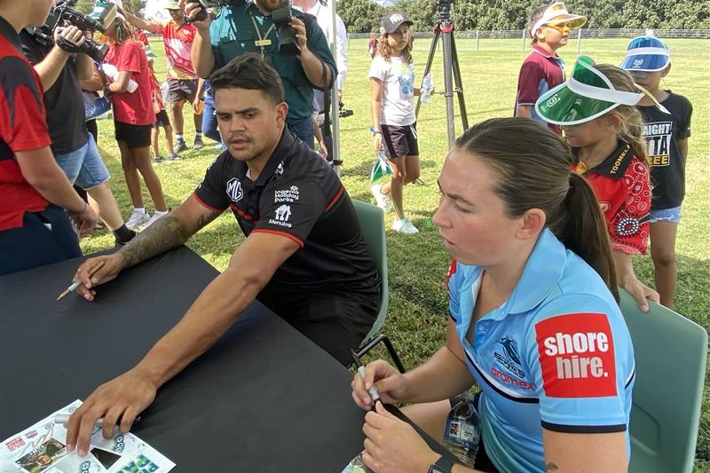A man and a woman sit at a table at a park, signing autographs.