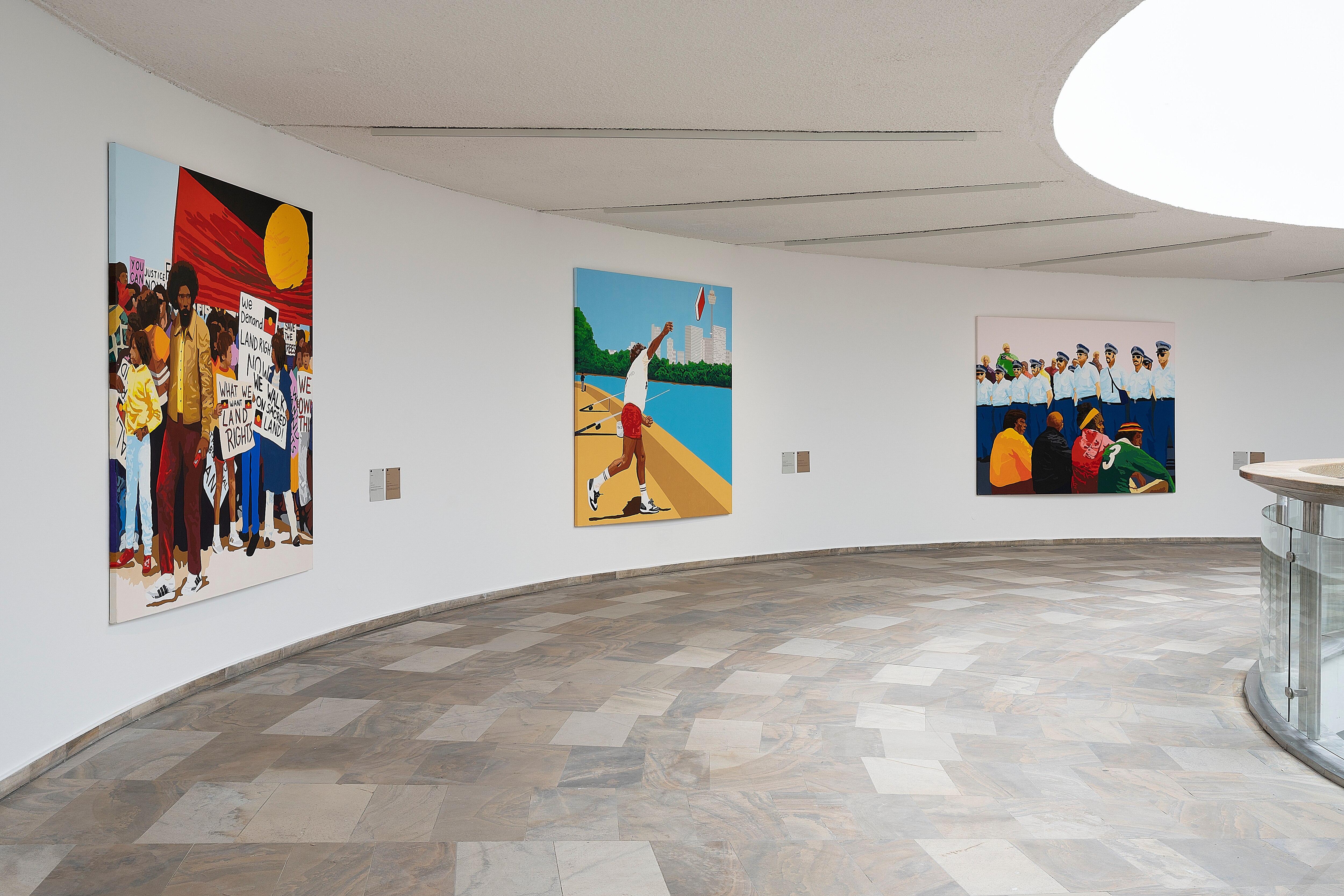 Three large canvas paintings hang on the white wall of a galley, each depicting Indigenous Australian subjects.